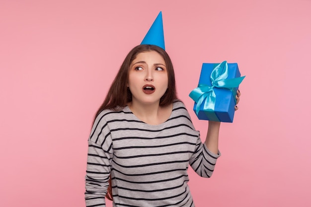 O que está dentro. Retrato de menina com chapéu de cone de festa segurando a caixa de presente perto da orelha e ouvindo, adivinhando a surpresa de aniversário, em antecipação ao presente interessante. tiro de estúdio interno, fundo rosa