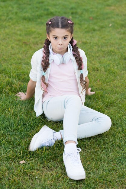 O que faz a criança feliz Penteado de tranças de menina e fones de ouvido modernos gostam de relaxar Segredos para criar uma criança feliz Menina linda criança fundo de grama verde Criança feliz emocional saudável relaxando ao ar livre