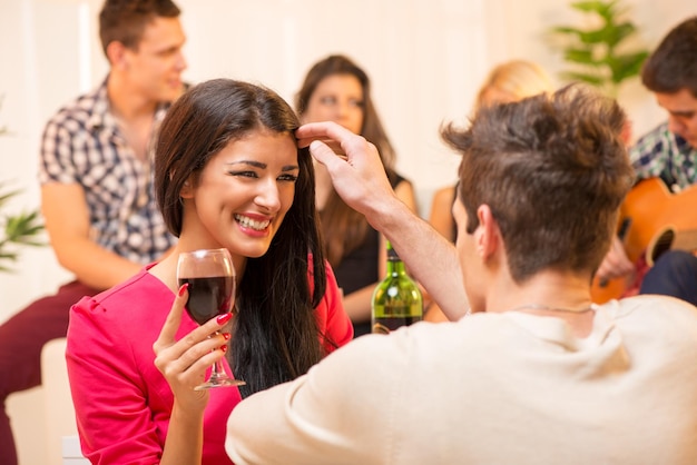 O rapaz da festa na casa está cortejando a linda morena acariciando seus cabelos, e ao fundo você pode ver um pequeno grupo de jovens se divertindo em uma festa.