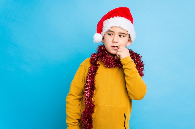 O rapaz pequeno que comemora o dia de Natal que veste um chapéu de Santa isolou o pensamento relaxado sobre algo que olha um espaço da cópia.
