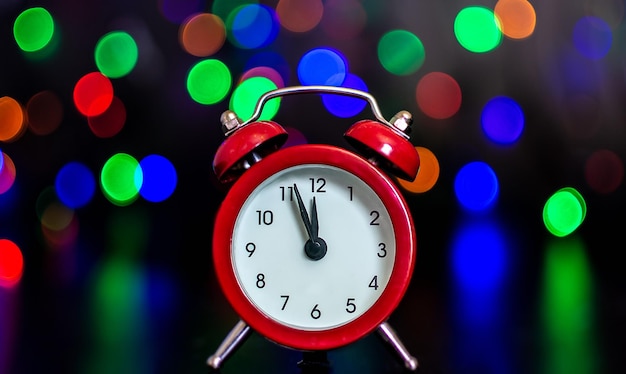 O relógio com ponteiros mostra cinco minutos para a meia-noite contra o fundo de luzes coloridas. Bokeh