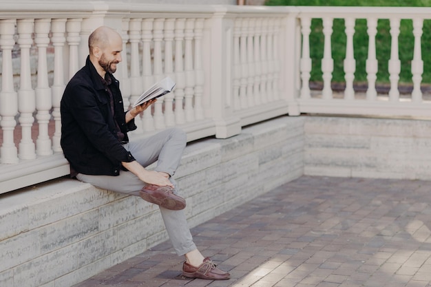 O retrato ao ar livre de um homem de meia-idade feliz com aparência atraente mantém o livro lê algo engraçado gosta de tempo livre em poses de parque contra o interior da varanda branca Pessoas e conceito de hobby