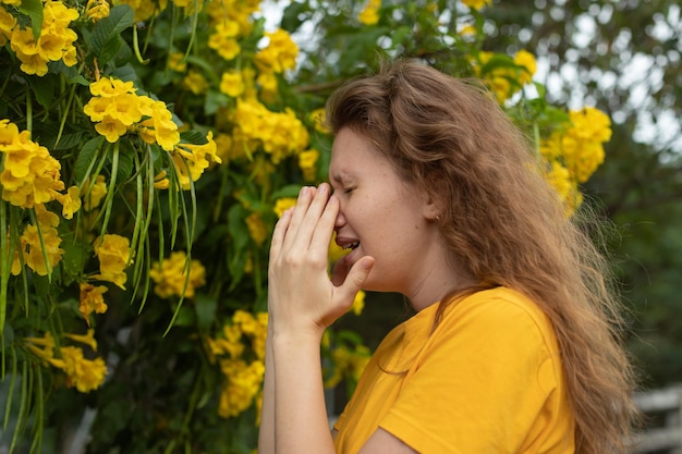 O retrato da bela jovem alérgica está sofrendo de alergia ao pólen ou frio no fundo da árvore florida natural na primavera ou no dia ensolarado de verão espirra soprando o nariz escorrendo esfrega os olhos