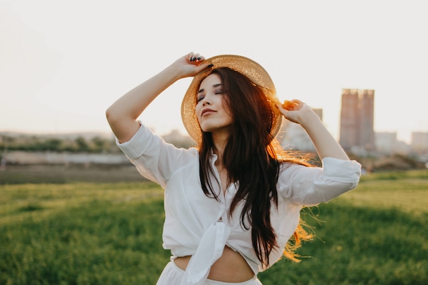 O retrato da menina asiática despreocupada bonita dos cabelos longos na roupa branca e no chapéu de palha aprecia a vida no campo