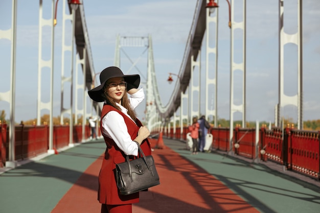 O retrato da moda da jovem modelo usa um traje vermelho, chapéu preto, óculos elegantes e bolsa. Espaço para texto