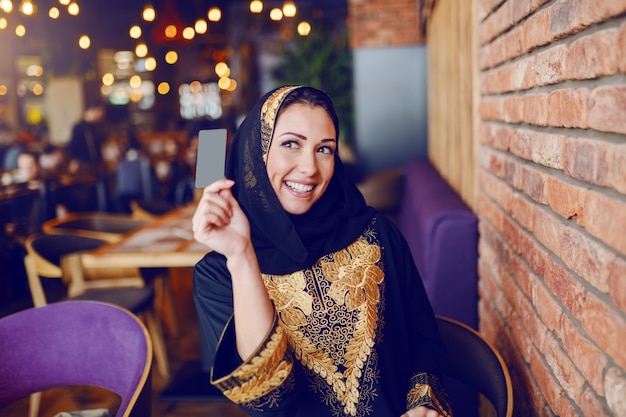 O retrato da mulher muçulmana lindo vestiu-se no desgaste tradicional que pede a verificação e que guarda o cartão de crédito ao sentar-se no café.
