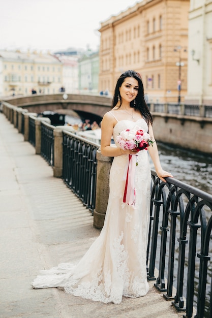 O retrato da noiva fêmea atrativa bonita veste o vestido de casamento branco longo, mantém o buquê