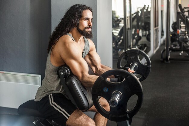 O retrato da vista lateral do jovem fisiculturista adulto com cabelos longos e encaracolados é treino na academia sozinho e se prepara para levantar a barra fazendo exercícios para o conceito de estilo de vida saudável interno de bíceps