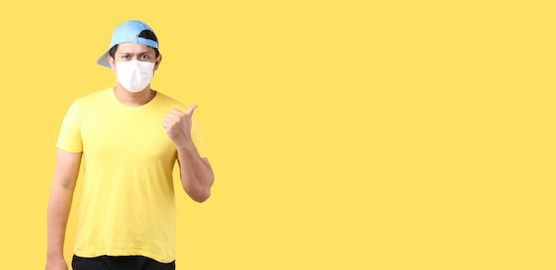 O retrato de chapéus asiáticos consideráveis do desgaste de homem e vestindo uma máscara está doente apontando o dedo isolado no fundo amarelo no estúdio com espaço da cópia.