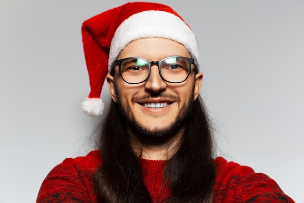 O retrato de estúdio de um jovem sorridente com cabelos longos e óculos usa suéter vermelho de Natal e chapéu de Papai Noel em um fundo branco