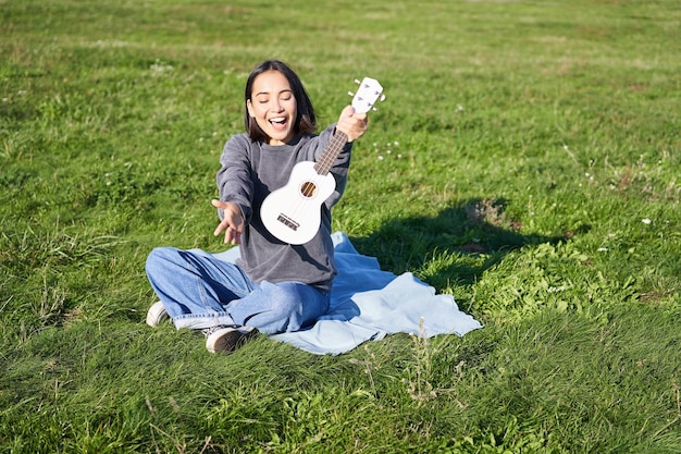 O retrato de música e instrumentos de uma linda garota asiática mostra seu ukulele branco tocando no parque enquanto está sentado