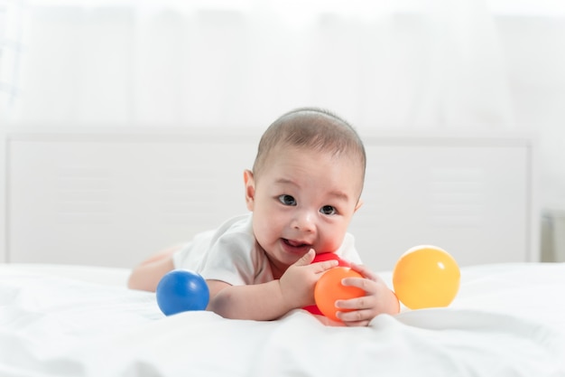 O retrato de um bebê de rastejamento na cama em seu quarto e jogando a bola brinca, bebê adorável no quarto ensolarado branco.