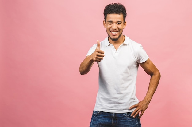 O retrato de um homem afro-americano novo de sorriso alegre vestiu-se em ocasional isolado contra o fundo cor-de-rosa, mostrando os polegares acima.