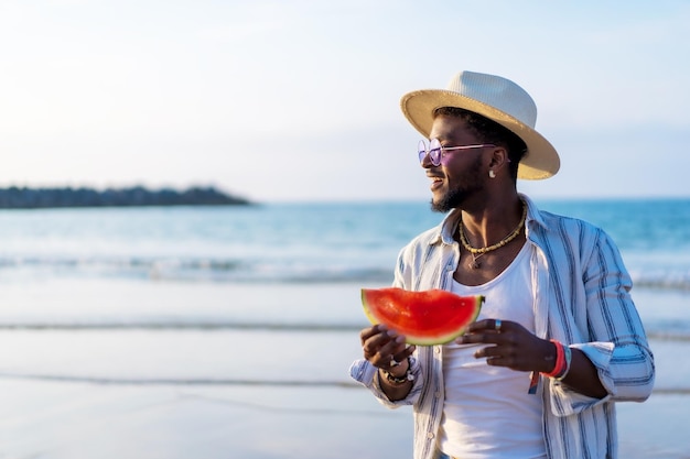 O retrato de um homem étnico negro aproveita as férias de verão na praia comendo uma melancia