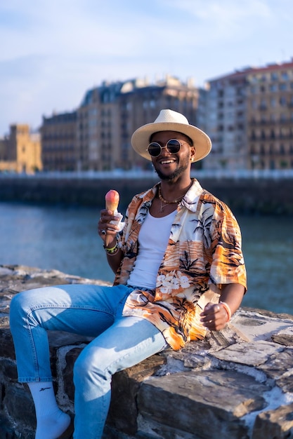 O retrato de um homem negro étnico aproveita as férias de verão na praia comendo um sorvete sentado à beira do rio