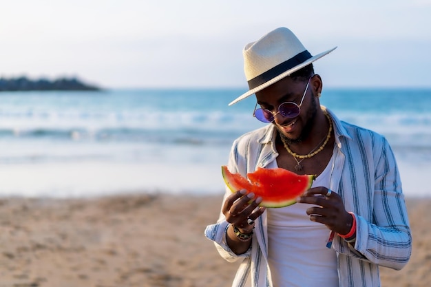 O retrato de um homem negro étnico aproveita as férias de verão na praia comendo uma melancia à beira-mar