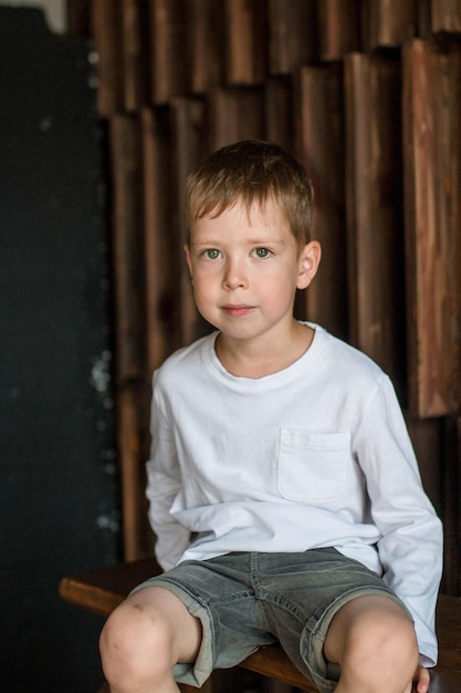 O retrato de um menino loiro em uma camiseta branca senta-se contra o fundo de uma parede de madeira e sorri olha para a câmera