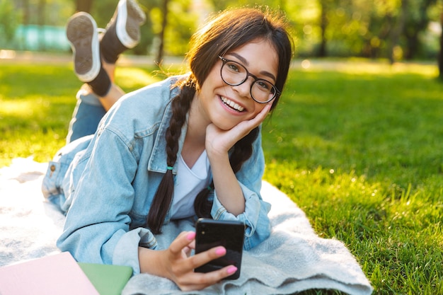 O retrato de uma alegre sorridente jovem estudante jovem usando óculos ao ar livre no parque natural usando telefone celular encontra-se na grama.