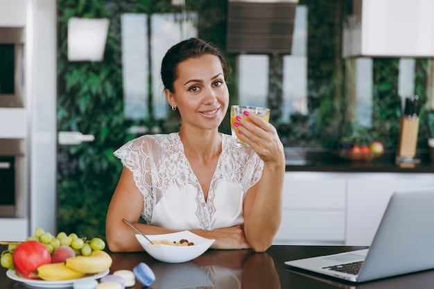 O retrato de uma bela jovem trabalhando com um laptop, enquanto o café da manhã com cereais e leite e bebendo suco de laranja.