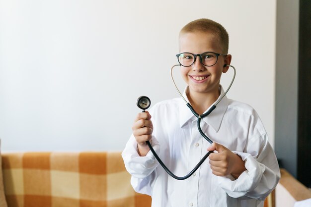 O retrato de uma criança do sexo masculino, caucasiano, em uma túnica de médicos com um estetoscópio na mão e óculos joga ...