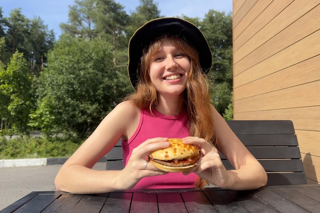 O retrato de uma jovem feliz está comendo um hambúrguer delicioso e suculento do lado de fora do café olhando para a câmera