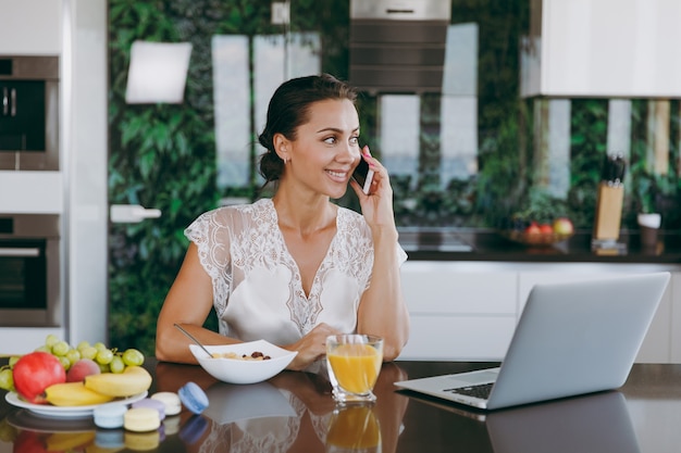 O retrato de uma linda mulher feliz falando em um telefone celular enquanto o café da manhã com o laptop na mesa