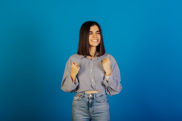 O retrato de uma mulher feliz e bonita com um sorriso largo levantou as mãos e comemorou a vitória, o sucesso isolado na superfície azul.