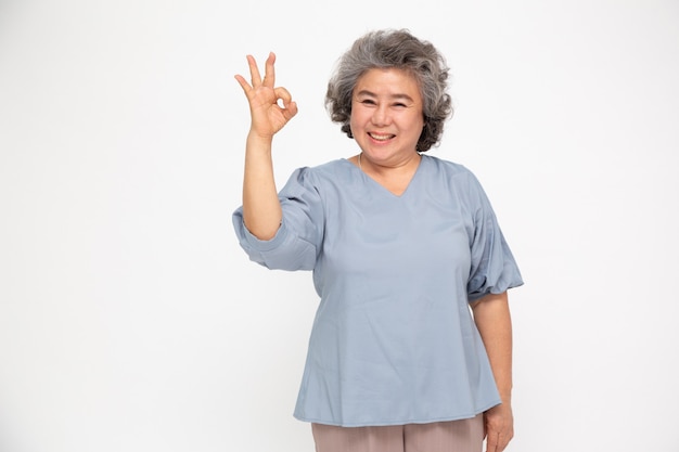 O retrato do gesto asiático sênior feliz da mulher ou a exibição da mão estão bem e olhando a câmera isolada na parede branca, sentimento mais velho da fêmea positivo e apreciam o conceito