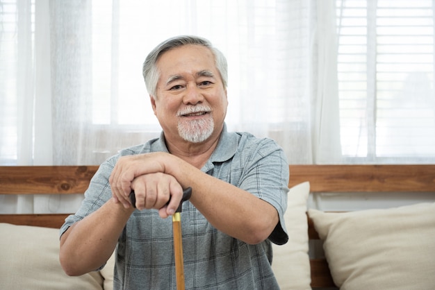 O retrato do homem asiático idoso sênior senta-se no olhar da bengala da preensão da mão do treinador na câmera com sorriso.