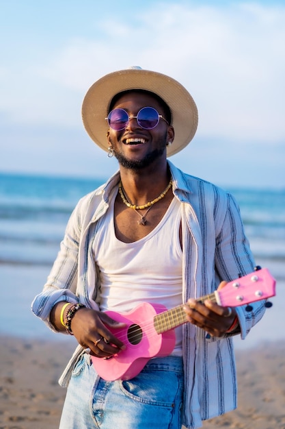 O retrato do homem negro étnico aproveita as férias de verão na praia tocando ukulele à beira-mar