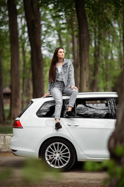 O retrato do modelo moreno bonito da forma "sexy" da mulher no revestimento das calças de brim que senta-se no carro branco luxuoso e relaxa. Conceito de moda. Idéia para sessão de fotos de garota com carro. Vertical.