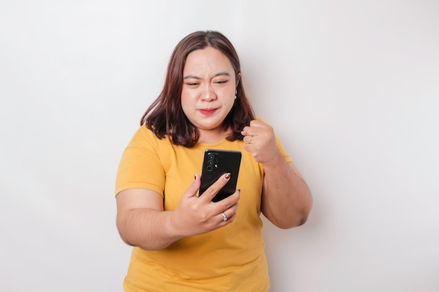 O rosto irritado e louco de uma mulher asiática de grande porte em camisa amarela enquanto segura seu telefone em fundo branco isolado