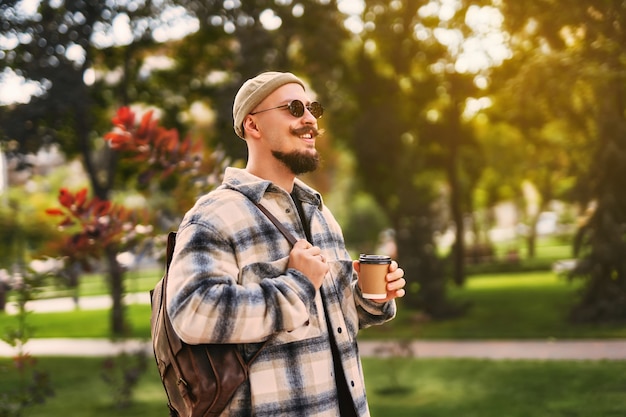 O rosto sorridente de um homem elegante e feliz passeando pelo parque ao ar livre com um café para viagem enquanto relaxa