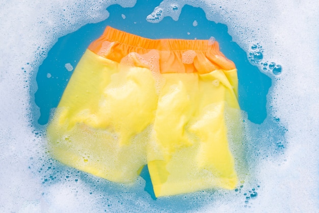O short amarelo e alaranjado do bebê embebe na dissolução da água do detergente para a roupa do bebê, pano de lavagem, conceito da lavanderia.