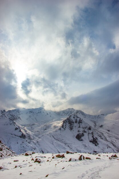 O sol ilumina montanhas de neve. Nuvens fecham o céu.