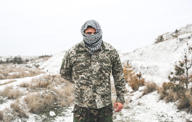 O soldado está de pé com o uniforme de camuflagem e a bandana xadrez keffiyeh shemagh. O homem está ao ar livre em um lugar abandonado e deserto.