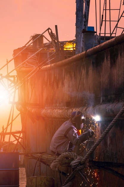 O soldador na plataforma está soldando a parede metálica do antigo navio de pesca na área do estaleiro na hora do pôr do sol