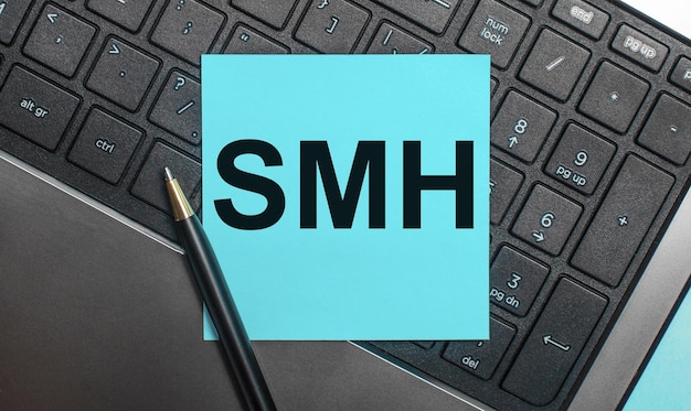 O teclado do computador possui uma caneta e um adesivo azul com o texto SMH. Postura plana.