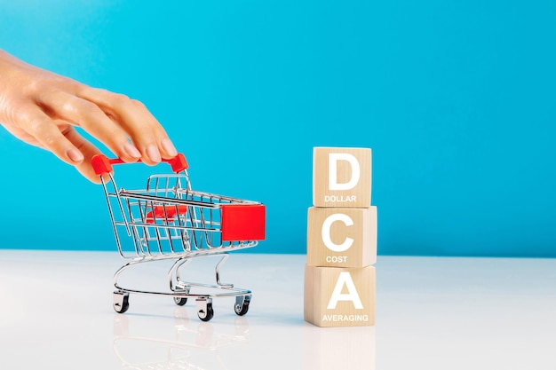 O texto DCA é escrito em cubos de madeira e a mão de uma mulher empurrando um carrinho de compras vazio no estúdio sobre fundo azul Conceito de negócios DCA abreviação de média de custo em dólar