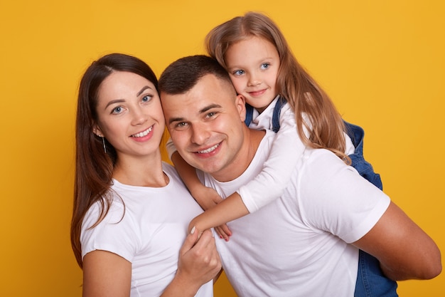 O tiro horizontal da família feliz que veste as camisas brancas, sorri do suporte isolado sobre o estúdio amarelo, pai que anda às cavalitas sua criança fêmea adorável. Conceito de relacionamento, felicidade e devoção.