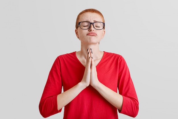 O tiro horizontal da mulher de cabelo vermelha esperançosa com olhos fechados, pressiona as palmas das mãos junto, suplicou a expressão, curva os lábios, implora por perdão, isolado no branco. A fêmea reza interna