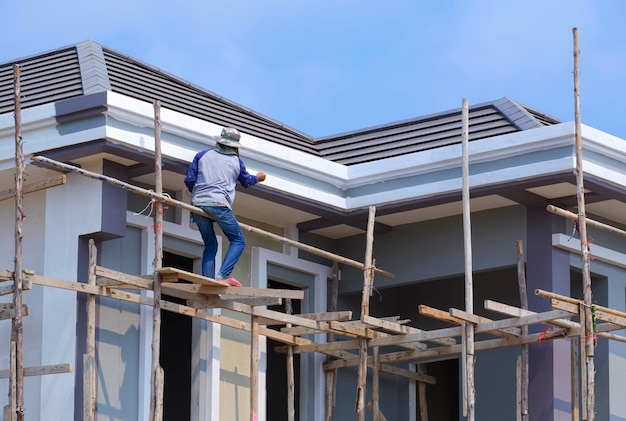 O trabalhador da construção civil no andaime de madeira está pintando os beirais do telhado da casa moderna contra o céu azul