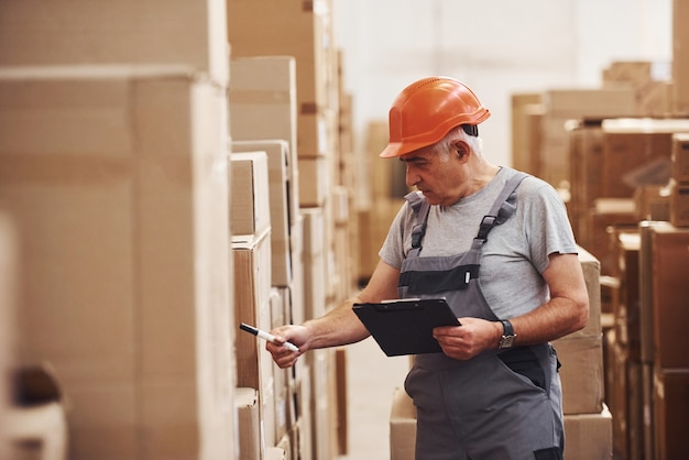 O trabalhador de armazenamento sênior de uniforme e bloco de notas nas mãos verifica a produção.