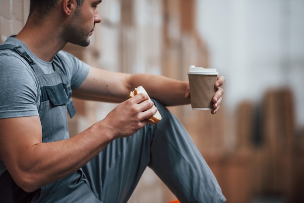 O trabalhador de armazenamento senta e faz uma pausa. Come sandwitch e bebe café.