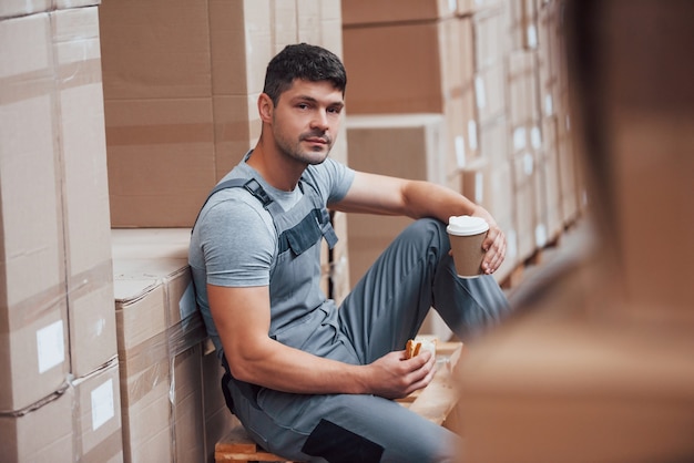 O trabalhador de armazenamento senta e faz uma pausa. Come sandwitch e bebe café.