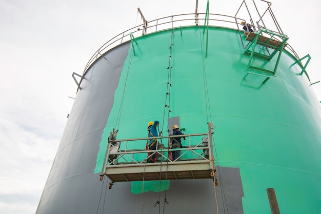 O trabalhador do sexo masculino leva o teleférico segurando um pincel de pintura industrial usado para superfície de superfície de óleo de tanque de placa de casca verde