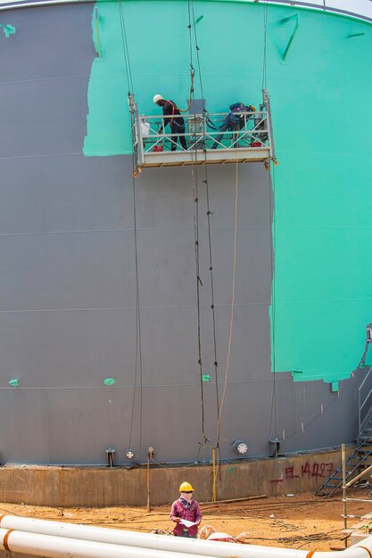 O trabalhador do sexo masculino leva o teleférico segurando uma escova de pintura industrial usada para a superfície da superfície do óleo do tanque da placa de concha cor verde