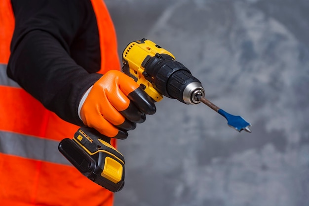 O trabalhador masculino segura uma chave de fenda elétrica sem fio em suas mãos contra o fundo de uma ferramenta de construção e uma parede de concreto