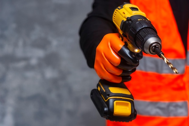 O trabalhador masculino segura uma chave de fenda elétrica sem fio em suas mãos contra o fundo de uma ferramenta de construção e uma parede de concreto
