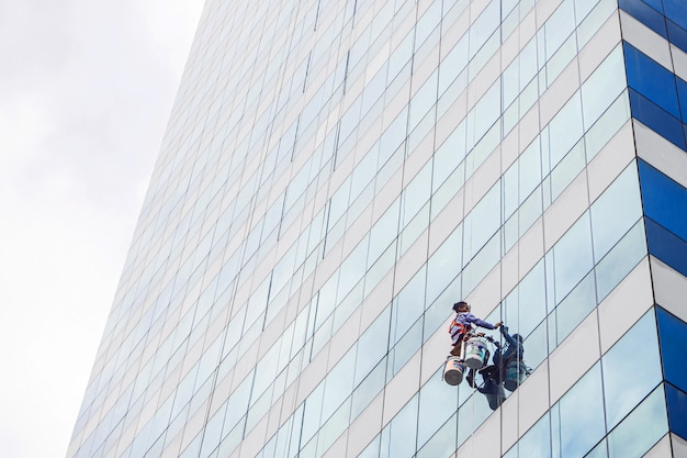 O trabalhador que limpa a janela de vidro no prédio alto com o alpinista de corda de suspensão.